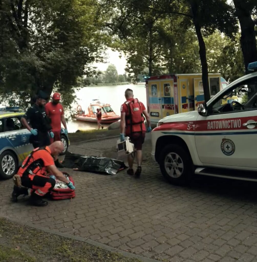 Ratownicy w czerwonych strojach przygotowują sprzęt medyczny obok wody, a w tle widoczne łodzie ratunkowe.