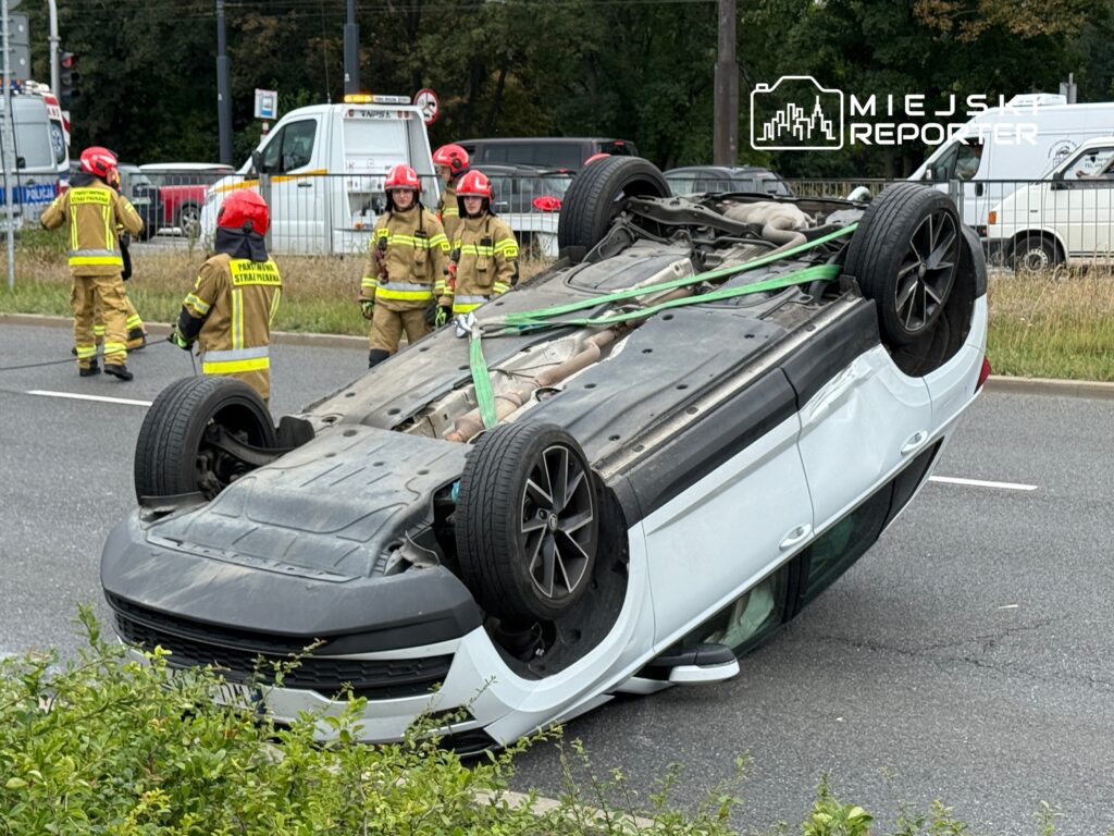 Kierowca w samochodzie osobowym leżącym na dachu, obok strażacy w mundurach pracujący przy akcji ratunkowej na drodze.