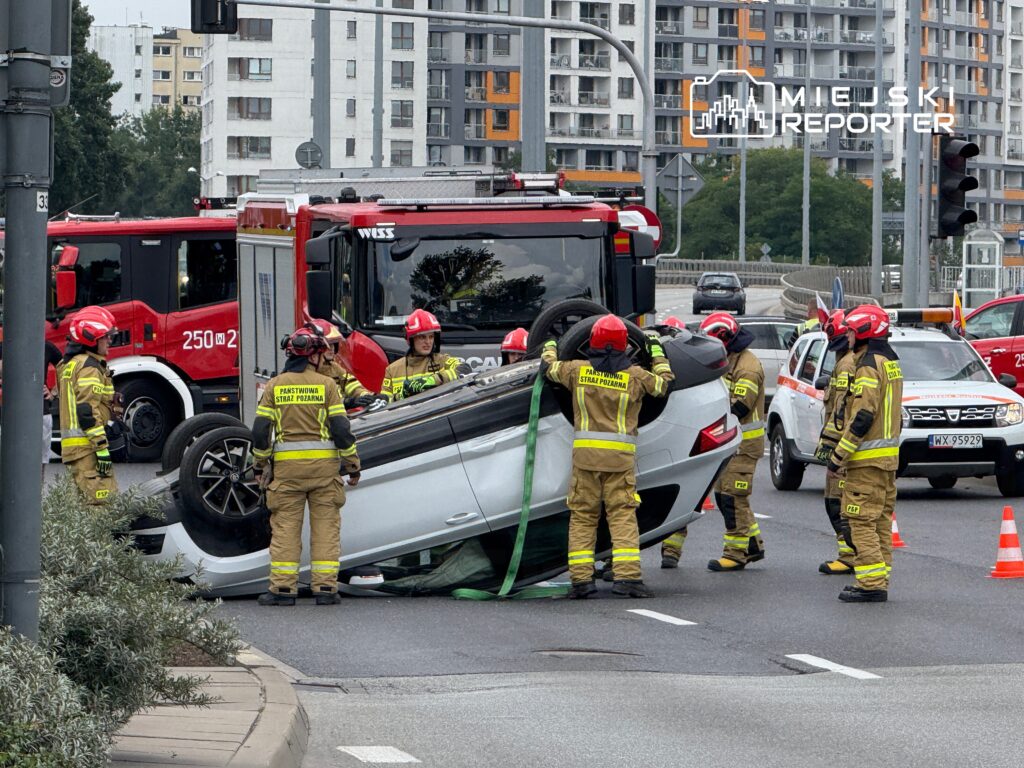 Grupa strażaków w mundurach próbuje postawić na koła przewrócone auto na ruchliwej drodze.