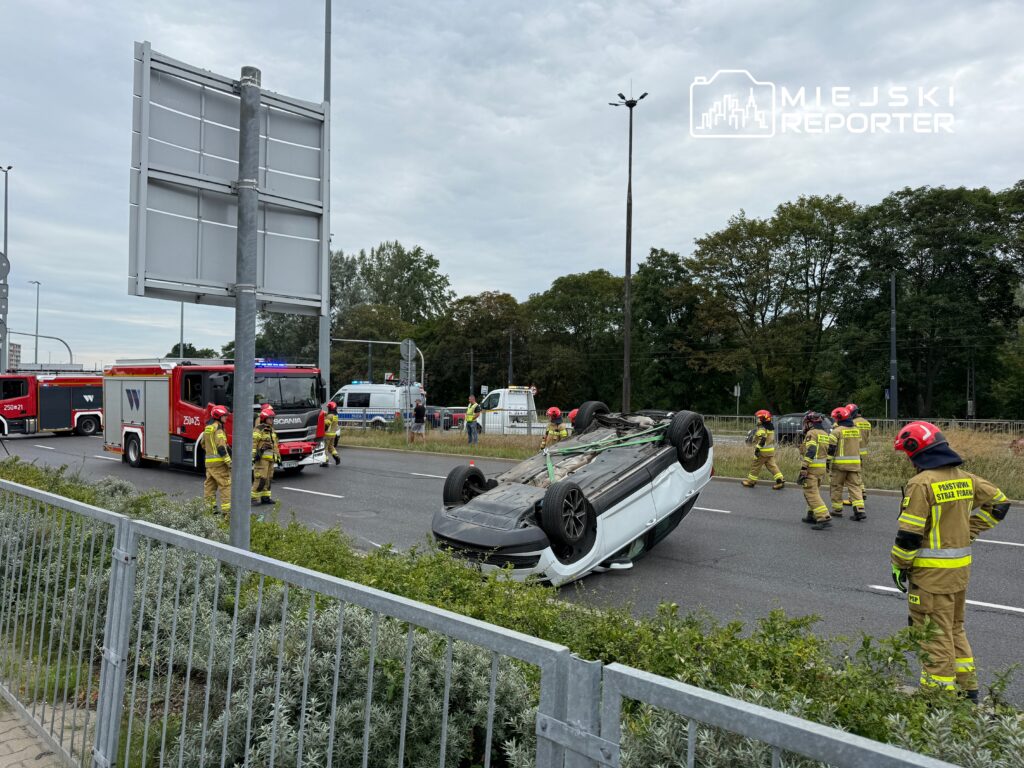 Na drodze leży przewrócony samochód, a w pobliżu grupa strażaków w hełmach ocenia sytuację.