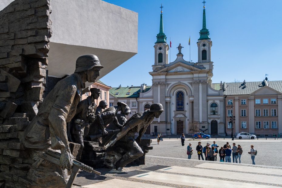 Grupa ludzi stoi na placu przed pomnikiem żołnierzy, w tle widoczny kościół z zielonymi wieżami.