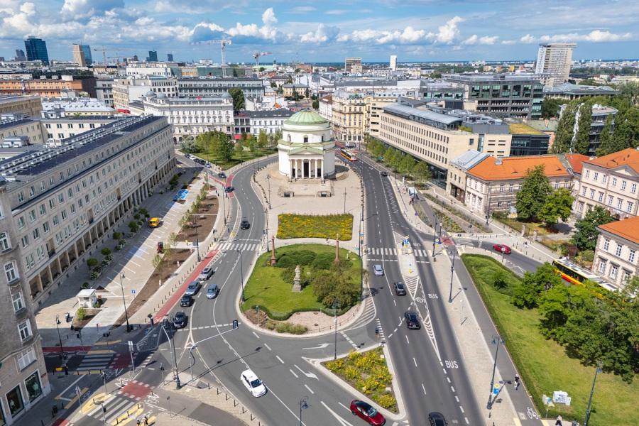 Widok na okrągły plac z zielonymi nasadzeniami, otoczony nowoczesnymi budynkami i ruchem ulicznym.