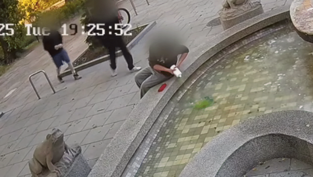 Mężczyzna w szarej koszulce siedzi na brzegu fontanny, trzymając w dłoniach biały przedmiot, podczas gdy dwie osoby przechodzą obok.