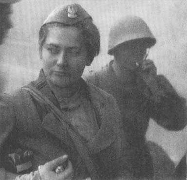 Młoda kobieta w mundurze wojskowym z odznaczeniami, stojąca w tle mężczyzny w hełmie, trzymającego papierosa.