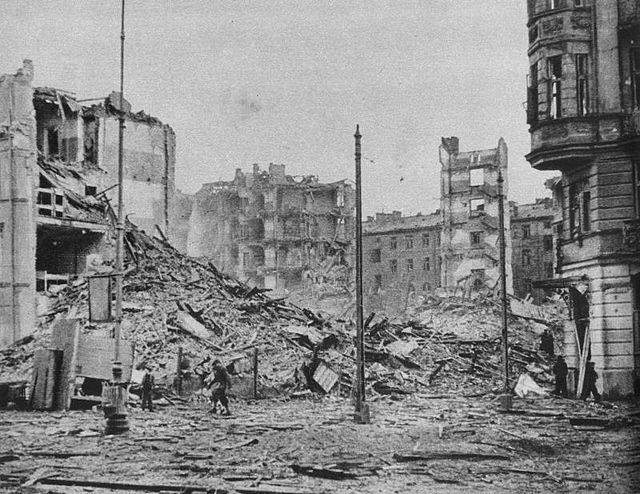 Zniszczony krajobraz miejski z gruzami budynków i fragmentami elewacji, w tle widoczni ludzie przeszukujący teren.