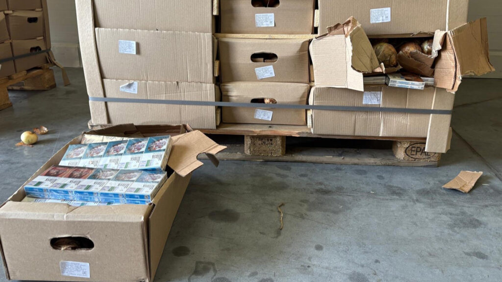 Kartony z produktami spożywczymi i papierosami na palecie w magazynie, z jednym otwartym kartonem.