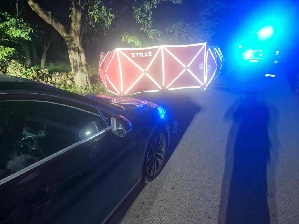 Na drodze leży przewrócony namiot z napisem "STRAŻ", obok stoi samochód z włączonymi sygnałami świetlnymi.