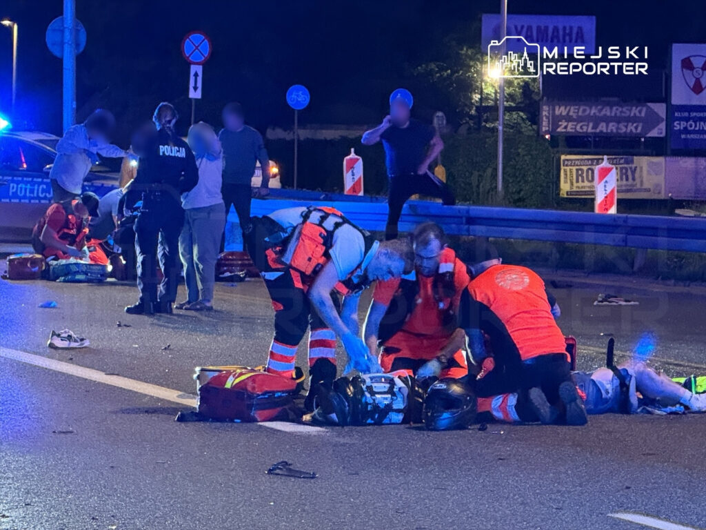 Ratownicy medyczni udzielają pomocy poszkodowanemu na drodze, w tle policyjny radiowóz i zebrani świadkowie.