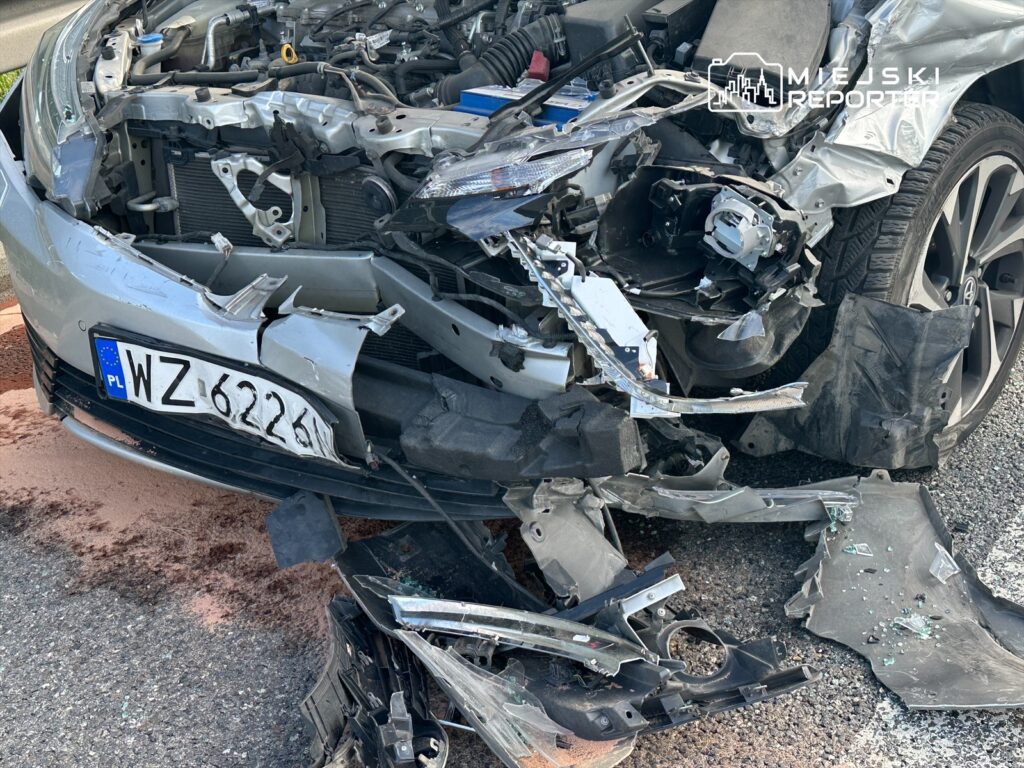 Zniszczony przód samochodu po wypadku drogowym. Widać uszkodzone elementy karoserii oraz rozbite lampy. Na jezdni leżą fragmenty pojazdu. Obraz ilustruje skutki kolizji, które mogą prowadzić do poważnych konsekwencji dla uczestników ruchu.