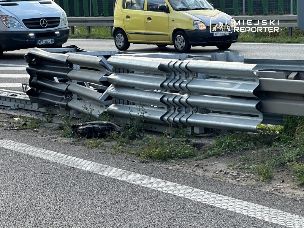 Uszkodzone bariery ochronne na drodze, obok przejeżdżających pojazdów. Widoczne zarośla oraz fragmenty nawierzchni. Sytuacja może stanowić zagrożenie dla bezpieczeństwa ruchu drogowego. W tle samochody osobowe, w tym żółty i szary. Obraz ilustruje problem infrastruktury drogowej.
