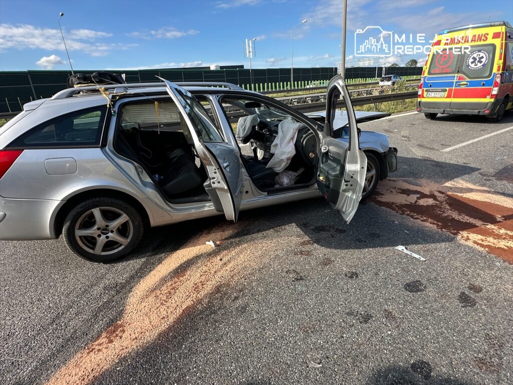 W wyniku wypadku drogowego na autostradzie zatrzymany został samochód osobowy marki Ford. Pojazd ma otwarte drzwi, a z wnętrza widać porozrzucane przedmioty. Na jezdni widoczny jest płyn, co sugeruje uszkodzenie. W tle widać ambulans, co wskazuje na interwencję służb ratunkowych.