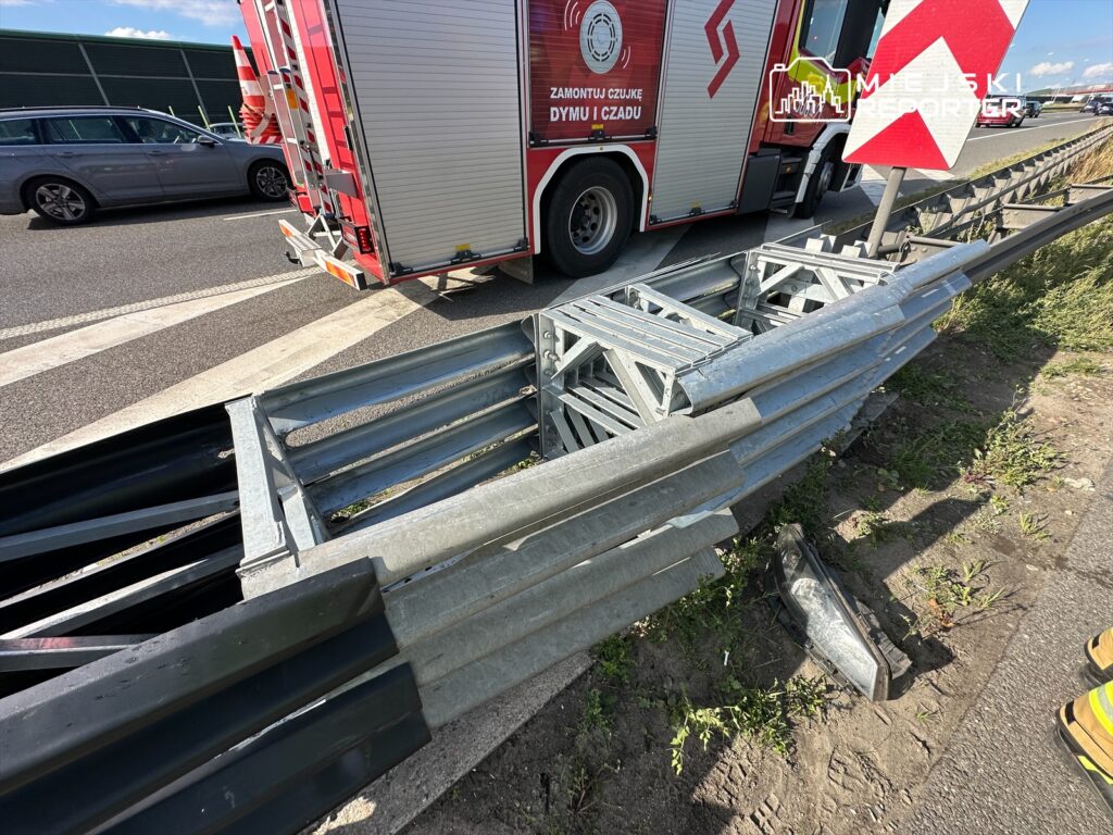 Zdjęcie przedstawia uszkodzone bariery ochronne na drodze, obok których stoi wóz strażacki. W tle widoczne są inne pojazdy. Sytuacja może sugerować zdarzenie drogowe, które wymagało interwencji służb ratunkowych. Na miejscu widać także oznakowanie drogowe.