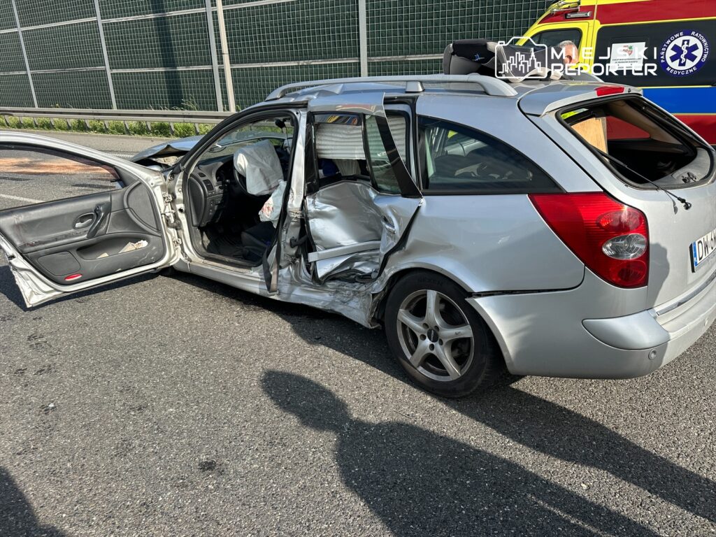 Zniszczony samochód osobowy na drodze, z otwartymi drzwiami i widocznymi uszkodzeniami bocznymi. Na tle widać karetkę pogotowia, co sugeruje, że miejsce to było sceną wypadku drogowego. Na jezdni widać ślady po zdarzeniu. Słońce oświetla miejsce, podkreślając powagę sytuacji.