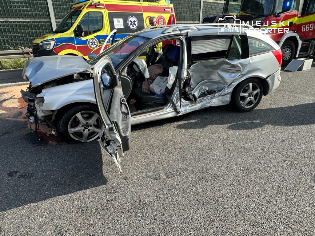 Wypadek drogowy z udziałem samochodu osobowego. Pojazd doznał poważnych uszkodzeń z lewej strony, a w środku widoczne są ślady krwi. W tle widać pojazd służb ratunkowych. Na miejscu zdarzenia pracują służby, które udzielają pomocy poszkodowanym.