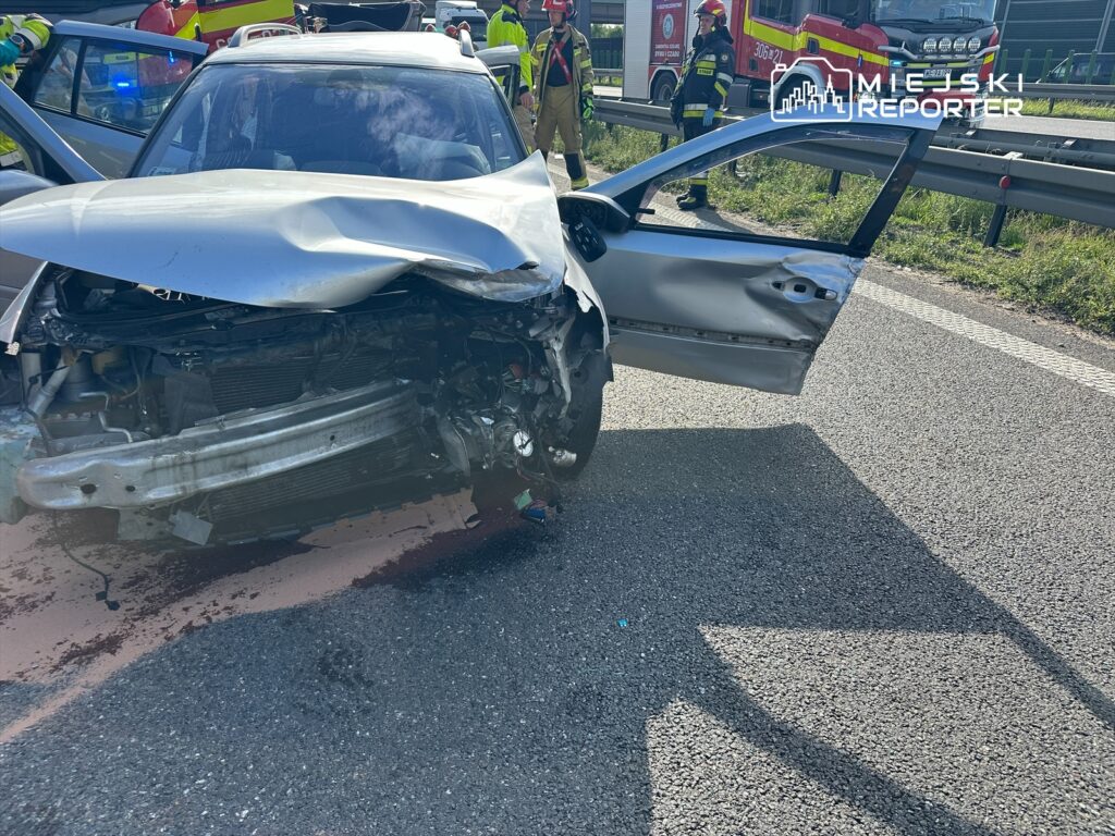 W wyniku wypadku drogowego uszkodzone zostało auto osobowe, które zatrzymało się na poboczu. Widoczne są zniszczenia karoserii oraz otwarte drzwi. Na miejscu zdarzenia pracują służby ratunkowe, w tym straż pożarna, zajmujące się zabezpieczeniem terenu.