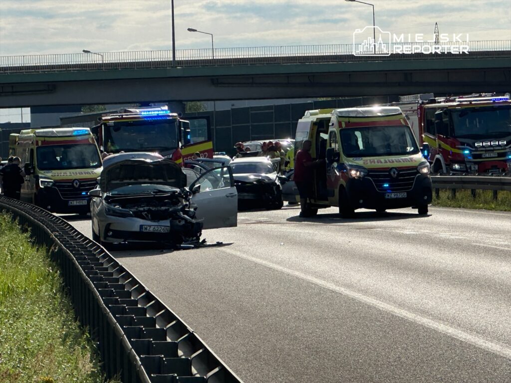 Wypadek drogowy na autostradzie. Uszkodzone pojazdy, w tym jeden z otwartą maską. Na miejscu obecne służby ratunkowe, w tym karetki i straż pożarna. Ruch wstrzymany, a na drodze widoczne są znaki ostrzegawcze. Sytuacja wymaga interwencji służb.