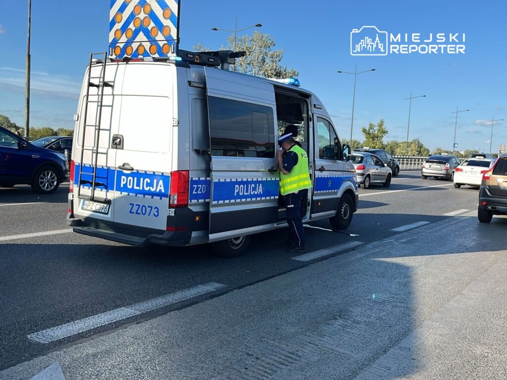 Policyjny radiowóz na drodze, z włączonymi sygnałami świetlnymi, zatrzymuje pojazdy w celu przeprowadzenia kontroli. Funkcjonariusz w kamizelce odblaskowej sprawdza dokumenty kierowcy. W tle widoczne są inne samochody poruszające się w kierunku przeciwnym. Słoneczna pogoda sprzyja intensywnemu ruchowi drogowemu.