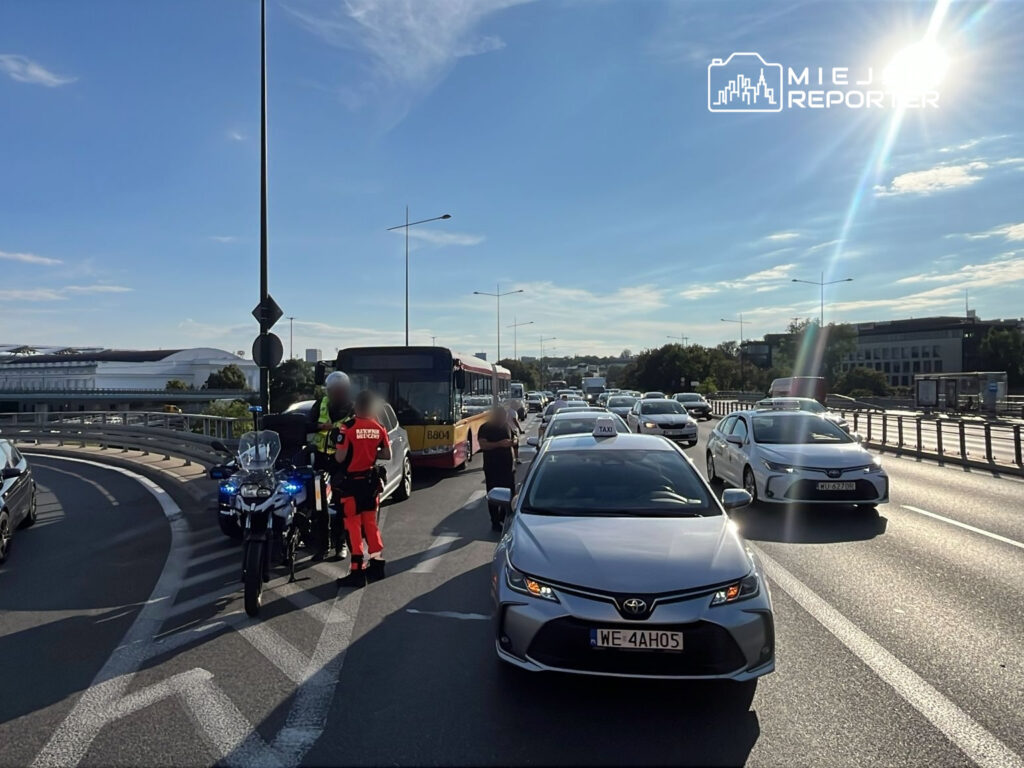 Na zdjęciu widać zator drogowy, w którym uczestniczy wiele pojazdów, w tym taksówki oraz autobusy. Policjant motocyklowy rozmawia z kierowcą, a w tle widoczne są budynki i znak drogowy. Słoneczna pogoda sprzyja widoczności. Zdarzenie miało miejsce na miejskiej drodze.