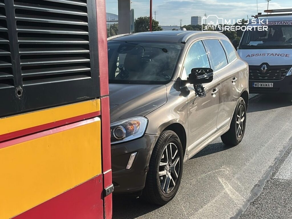 Zdarzenie drogowe w mieście: na ulicy widoczny jest zderzak autobusu oraz osobowy samochód w kolorze szarym. W tle znajduje się pojazd służb pomocowych. Na jezdni widać ślady opon. Sytuacja mogła wpłynąć na ruch drogowy w okolicy.