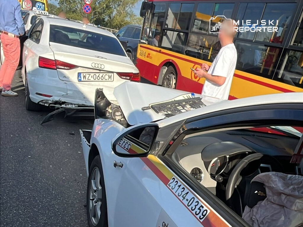 W wyniku kolizji drogowej doszło do uszkodzenia dwóch samochodów osobowych. W tle widoczny jest autobus komunikacji miejskiej. Na miejscu zdarzenia przebywają kierowcy oraz świadkowie. Służby porządkowe prowadzą działania mające na celu zabezpieczenie miejsca wypadku.