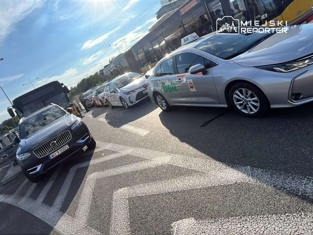 Zdj. przedstawia ruch drogowy w mieście, gdzie widoczne są różne pojazdy, w tym taksówki i autobus. W tle widać sygnalizację oraz kierowców czekających na możliwość włączenia się do ruchu. Słoneczna pogoda sprzyja intensywnemu ruchowi ulicznemu.