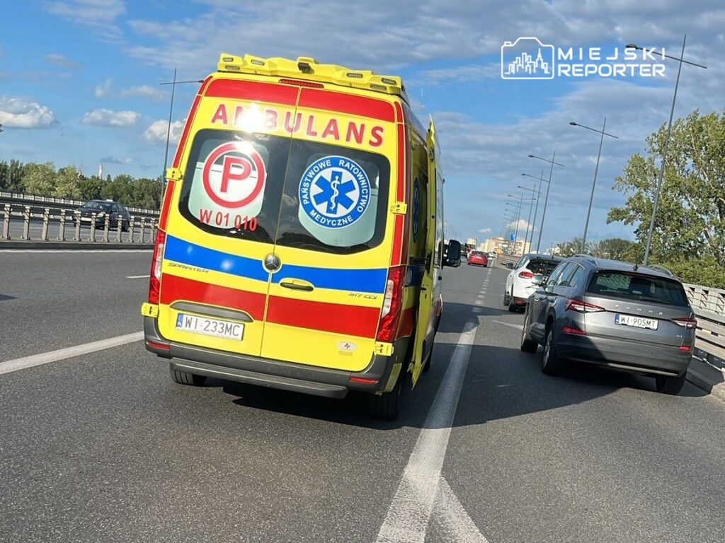 Ambulans z logo Państwowego Ratownictwa Medycznego porusza się po ruchliwej drodze. W tle widać inne pojazdy oraz chmurki na niebie. Na samochodzie widoczny jest znak zakazu parkowania. Obraz ilustruje sytuację na drodze, gdzie służby ratunkowe są w akcji.