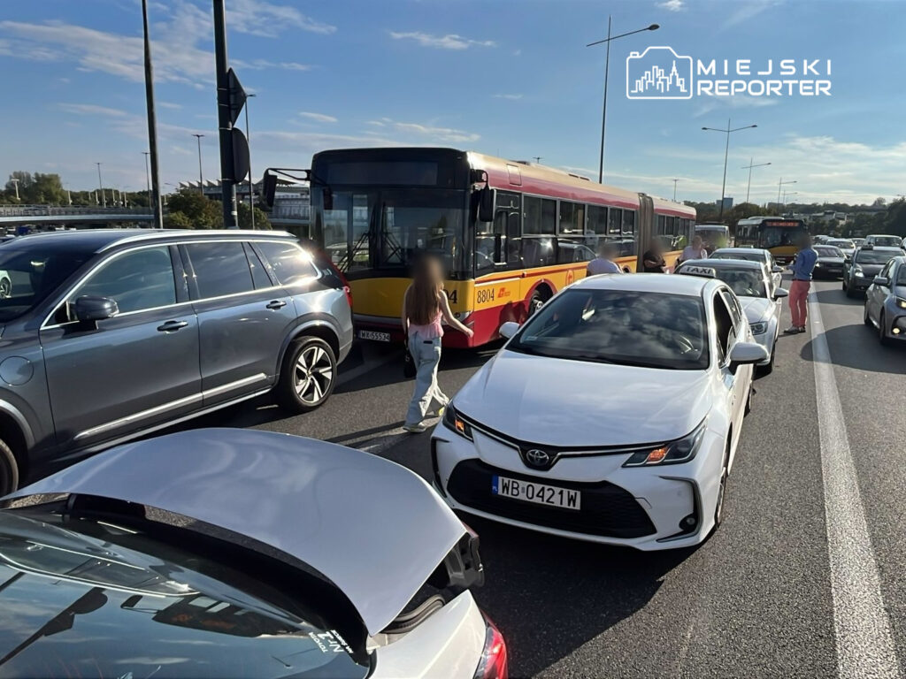 Zdarzenie drogowe na ruchliwej ulicy. Zablokowany ruch samochodowy, widać stojące pojazdy, w tym autobus i taksówkę. Na jezdni znajdują się piesi, którzy próbują przejść obok zatoru. Słońce świeci, a na niebie widoczne są chmury. Sytuacja może powodować opóźnienia w komunikacji.