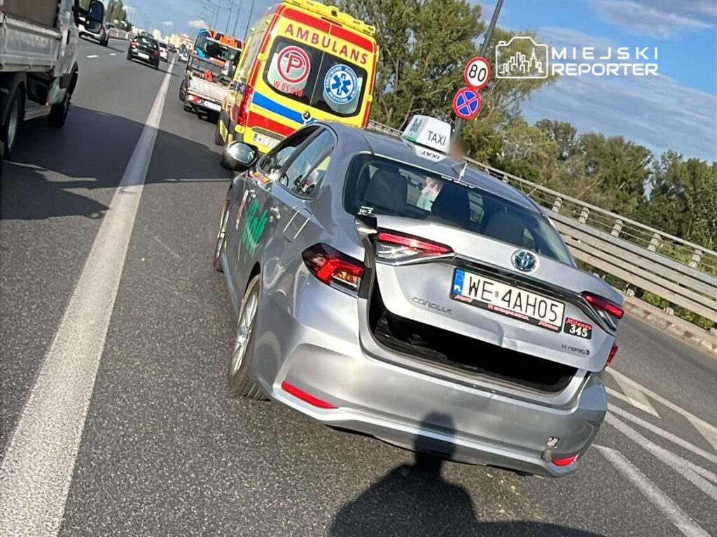 Na zdjęciu widoczny jest srebrny samochód osobowy typu taxi z widocznym oznaczeniem. W tle znajduje się pojazd ratunkowy oraz inne samochody poruszające się po drodze. Scena ma miejsce na ruchliwej jezdni, co sugeruje intensywny ruch drogowy. Znak ograniczenia prędkości wskazuje 80 km/h.