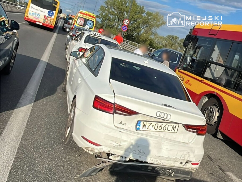 Zdarzenie drogowe na ruchliwej ulicy. W tle widoczne zablokowane pojazdy, w tym białe Audi A3 z uszkodzeniami. Na drodze obecne są także autobusy i taksówki. W okolicy przebywają osoby, które mogą być świadkami wypadku. Sytuacja spowodowała utrudnienia w ruchu.