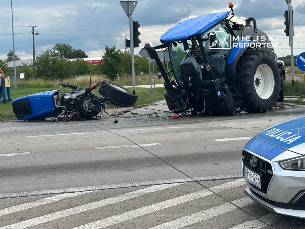 Traktor leży na boku na drodze, obok widać policyjny radiowóz oraz uszkodzone elementy maszyny.