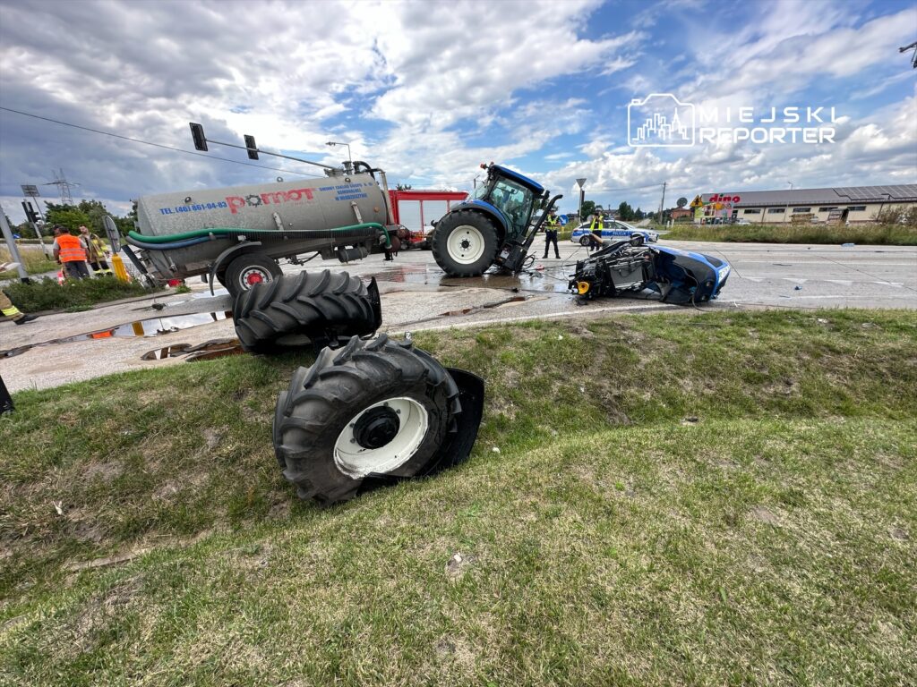 Traktor przewrócony na drodze, obok rozbite auto; w tle strażacy oraz pojazd gaśniczy.