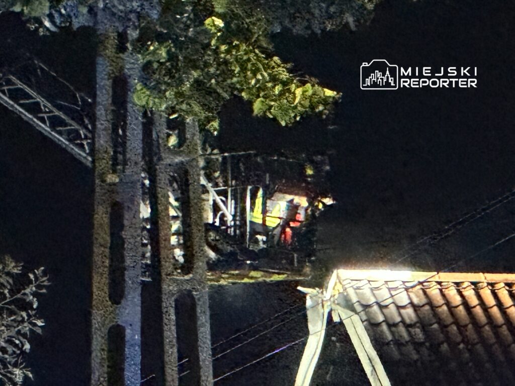 Wysoka drabina strażacka z włączonymi światłami, z której widać sylwetki strażaków, w nocy nad dachem budynku.