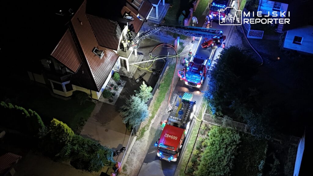 Strażacy na dachu budynku używają drabiny, podczas gdy w tle stoją wozy strażackie z włączonymi sygnałami świetlnymi.