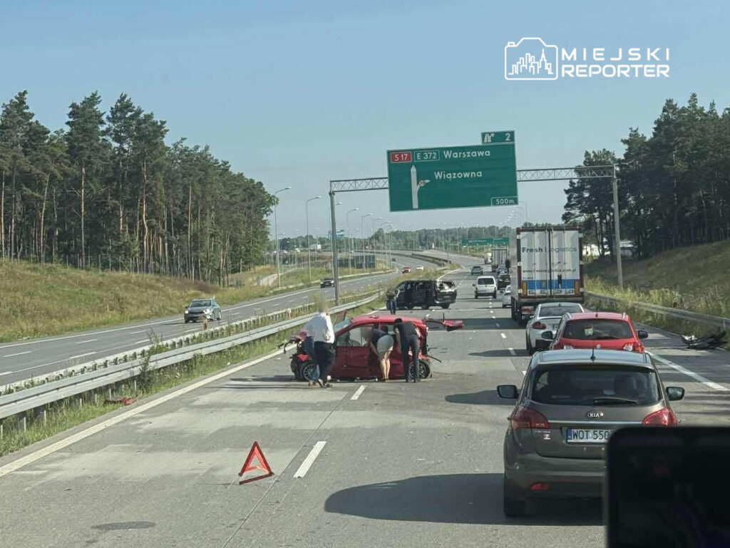 Na drodze ekspresowej widać wypadek, z udziałem czerwonego samochodu, obok którego stoją dwie osoby. W tle ruch pojazdów oraz znaki drogowe.