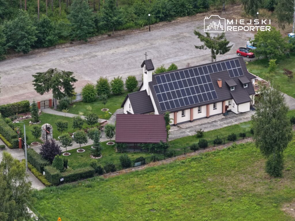 Kościół z panelami słonecznymi otoczony zielonymi drzewami i zadbaną przestrzenią zieloną. W tle widoczny parking.