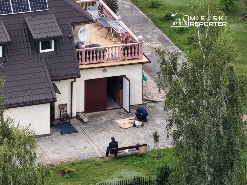 Mężczyzna w garniturze siedzi na ławce przed domem, podczas gdy inny mężczyzna pracuje przy materiałach budowlanych.