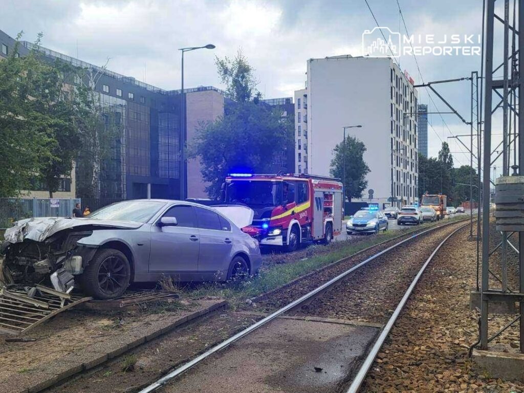 Uszkodzony samochód osobowy stoi na poboczu torów kolejowych, obok zjeżdżają pojazdy służb ratunkowych.