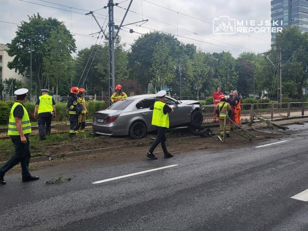 Grupa ratowników i policjantów pracuje przy uszkodzonym samochodzie na poboczu drogi, obok torów tramwajowych.