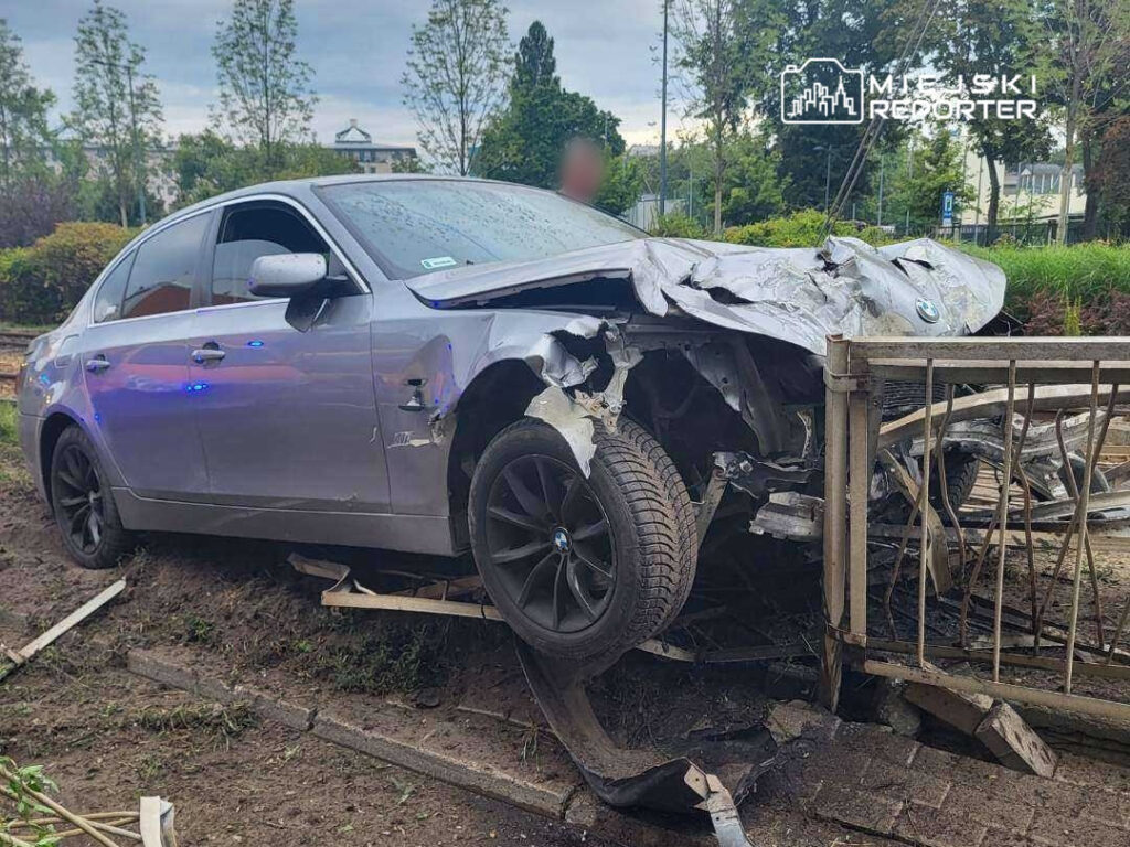 Uszkodzony samochód osobowy marki BMW wjeżdża w ogrodzenie, leżąc na stoku w otoczeniu zieleni.