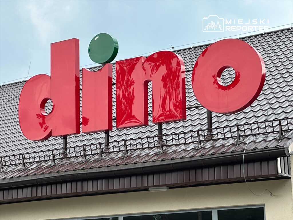 Wysoki, czerwony szyld z napisem "dino" z zieloną kropką na dachu budynku z dachówką.