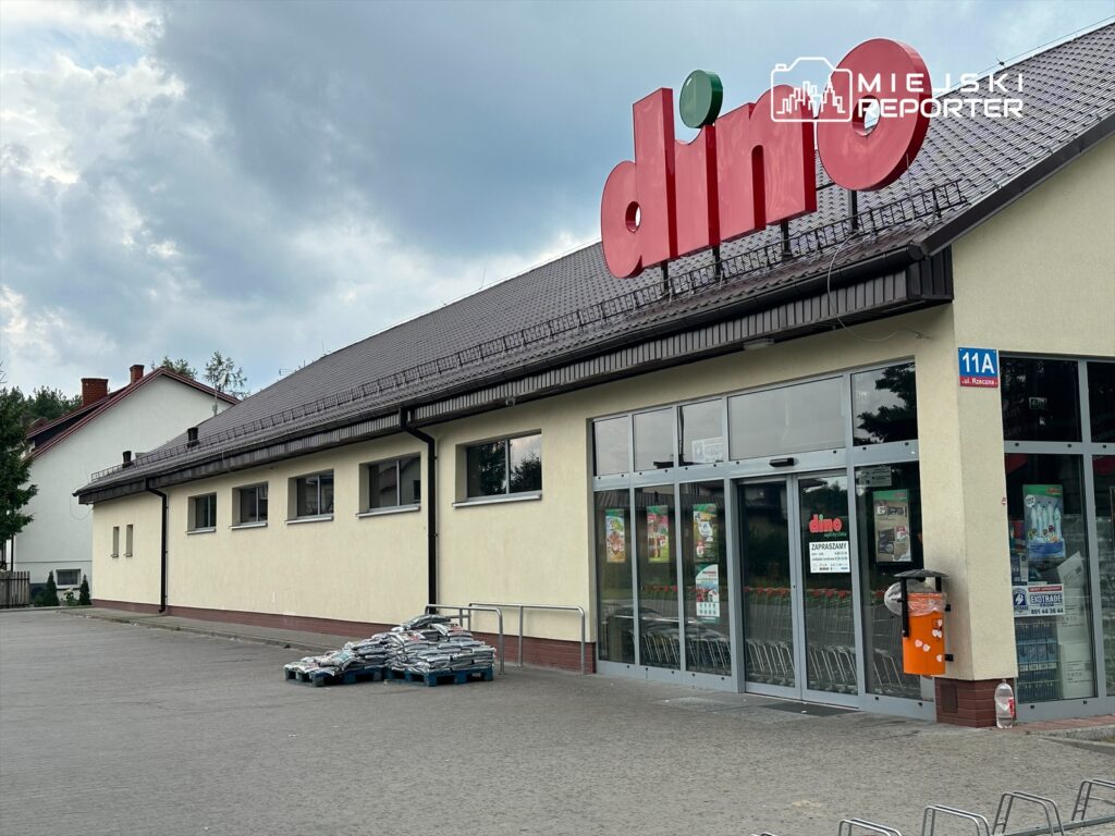 Sklep spożywczy z czerwonym logo "dino" i szklanymi drzwiami, obok paleta towarów na parkingu.