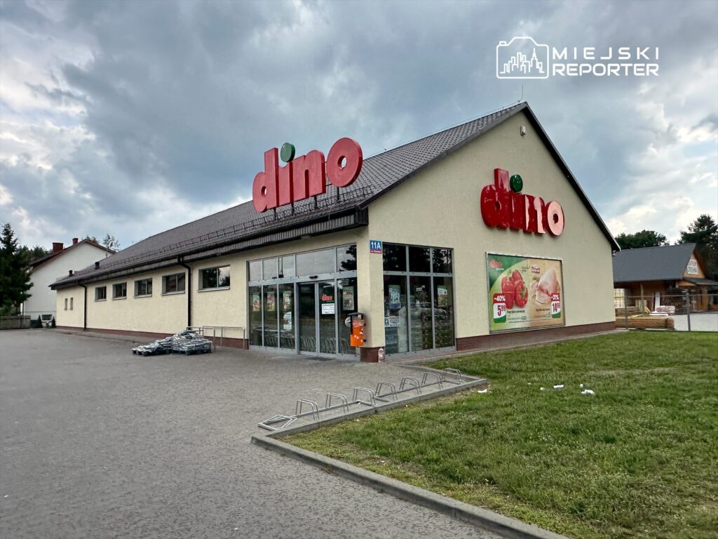 Supermarket „Dino” z dużym, kolorowym logo, z przeszklonymi drzwiami i reklamami na elewacji, w otoczeniu parkingu.