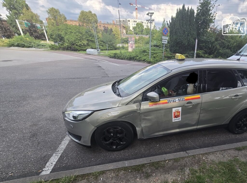 Złoty taksówkarz zaparkowany na poboczu, z uszkodzoną lewą stroną, obok pojazdu pomocy drogowej. W tle zieleń i budynki.