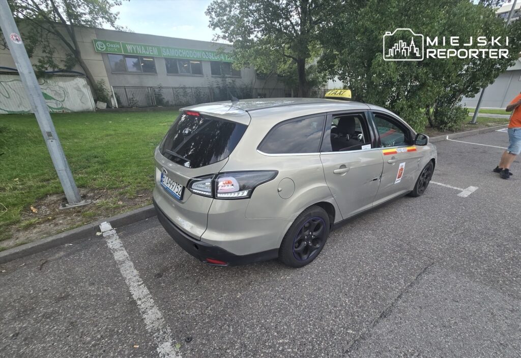 Taksi zaparkowane na parkingu obok budynku z napisem "WYNAJEM SAMOCHODÓW". W tle mężczyzna w pomarańczowej koszulce idzie w kierunku pojazdu.