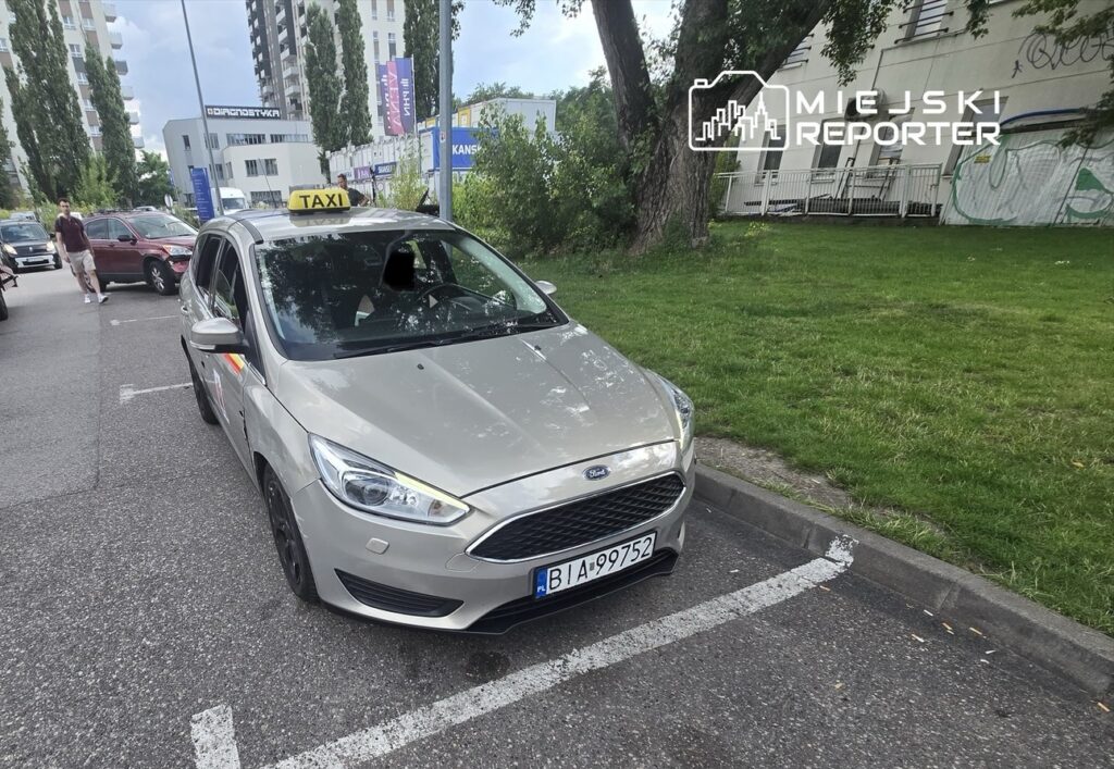 Taxi zaparkowane na poboczu drogi, obok mężczyzny w pomarańczowej koszulce, który rozmawia z kierowcą lawety.