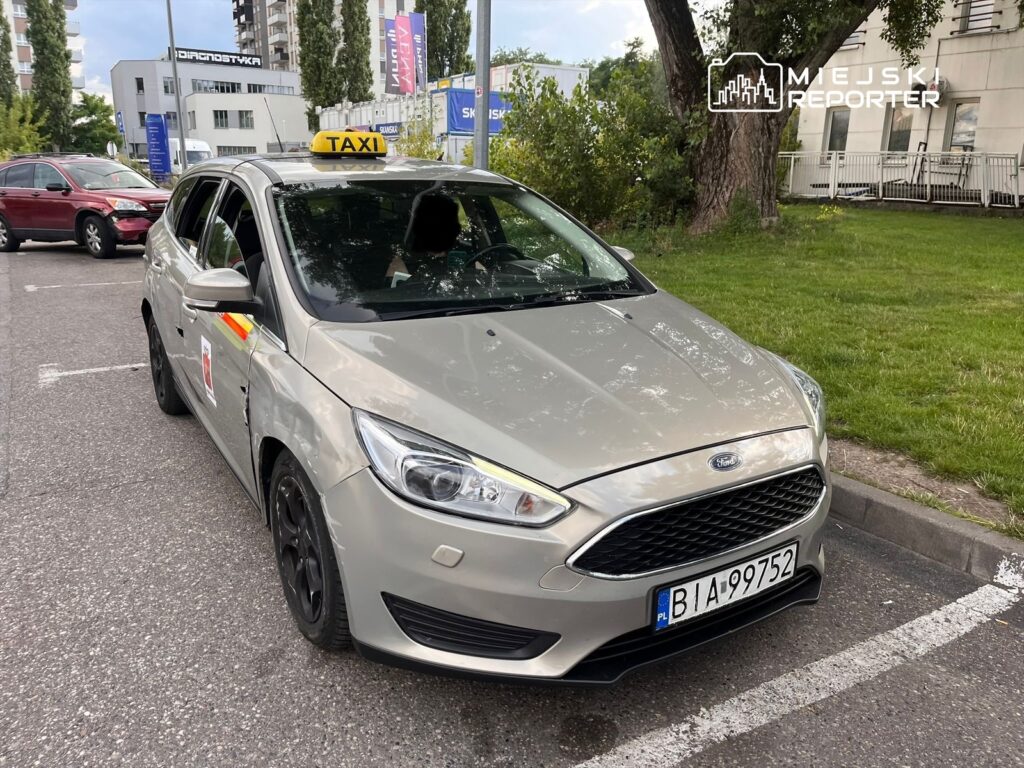 Taksi Ford stojące na parkingu, z widocznym znakiem "TAXI" na dachu i uszkodzeniami na boku.