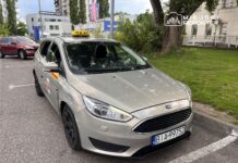 Taksi Ford stojące na parkingu, z widocznym znakiem "TAXI" na dachu i uszkodzeniami na boku.