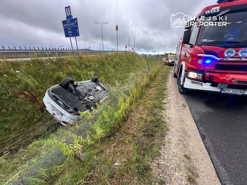 Na poboczu drogi leży przewrócone na dach białe auto, obok niego stoi wóz strażacki z włączonymi sygnałami.