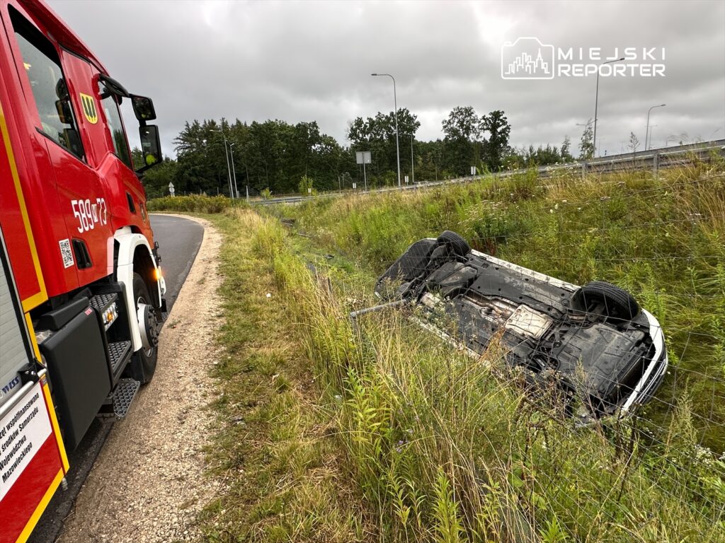 Czerwony wóz strażacki stoi na zakręcie drogi obok przewróconego samochodu w rowie, otoczonego wysoką trawą.