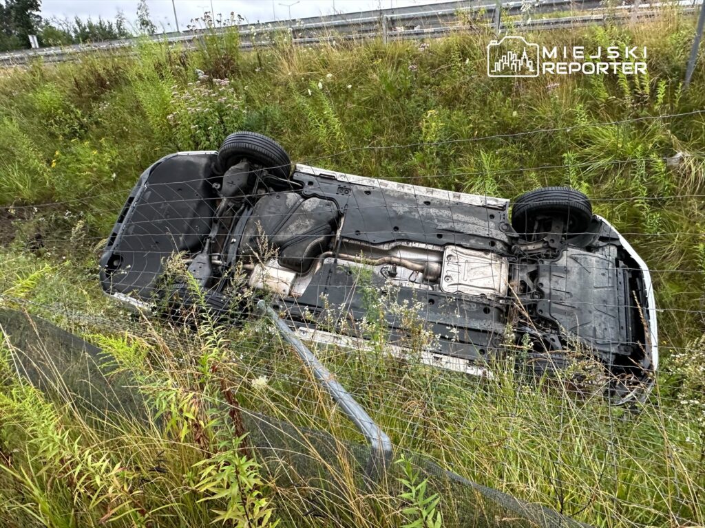 Przewrócony samochód leży na boku w wysokiej trawie przy drodze, z widocznym uszkodzeniem podwozia.